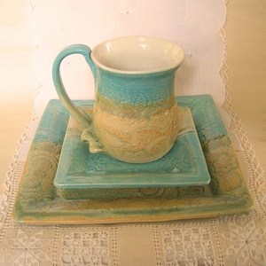 May include: A set of three ceramic plates in shades of teal, green, and yellow with a floral design. The set includes a mug with a matching design.