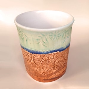 May include: A ceramic mug with a white interior and a light blue and brown exterior. The mug has a decorative design of leaves and flowers.