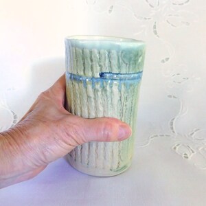 May include: A ceramic cup with a textured, striped design in shades of green, blue, and white. The cup has a glazed finish and a rounded shape.