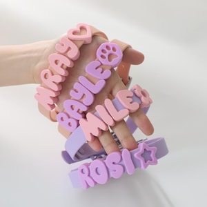 Puede incluir: Una colección de collares para mascotas en colores pastel con nombres escritos en letras grandes y de burbujas. Los nombres incluyen "WRAY", "BAYLE", "MILE" y "ROSI". Cada collar presenta un encanto diferente, como un corazón, una huella de pata o una estrella.