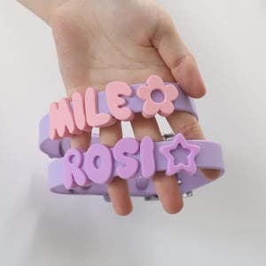 May include: Two light purple silicone collars featuring raised, bubble-style lettering. The top collar displays the name MILE in pink with a pink flower icon. The bottom collar displays the name ROSI in purple with a purple star icon.