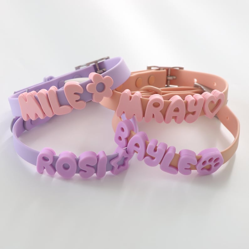 May include: Two silicone collars in lavender and peach, each featuring raised, block-letter names. The lavender collar displays the names MILE and ROSI with a flower and star icon. The peach collar shows MRAY and BAYLE with a heart and paw print.