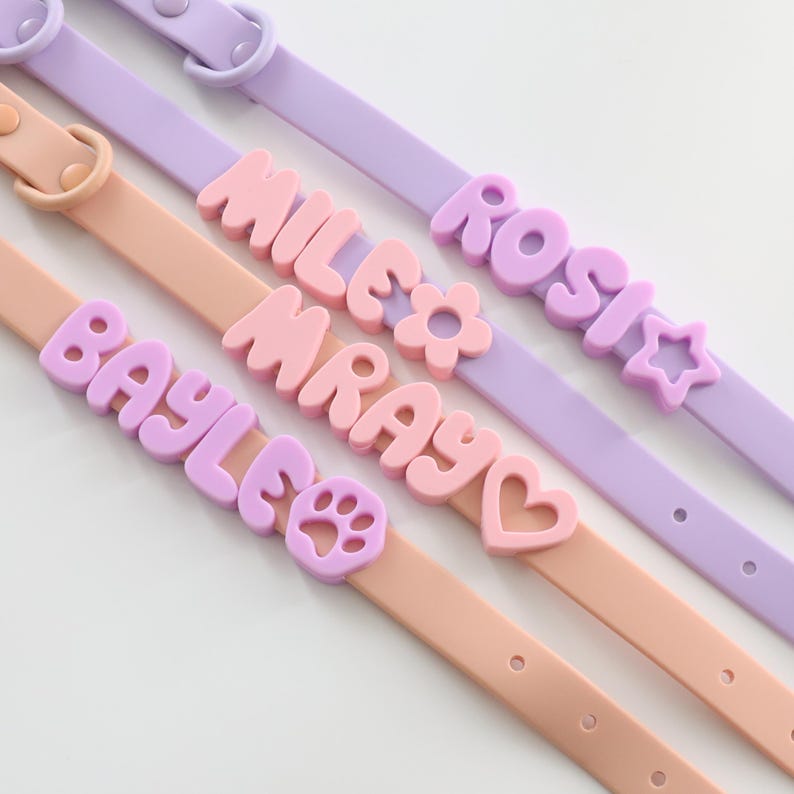 May include: Three silicone pet collars in light purple and peach colors. Each collar features raised, block-letter names including ROSI, MILE, MRAY, and BAYLE, accompanied by decorative charms shaped like a star, a flower, a heart, and a paw print.