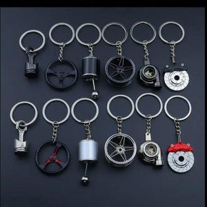 Puede incluir: Una colección de llaveros con temática automovilística. Los llaveros presentan réplicas en miniatura de piezas de automóviles, como pistones, volantes, ruedas y pinzas de freno. Los llaveros son plateados, negros y rojos, sobre un fondo oscuro.