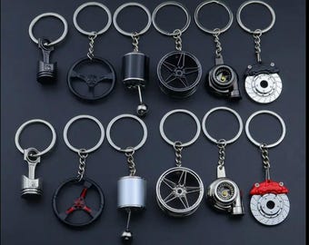 May include: A collection of automotive-themed keychains. The keychains feature miniature replicas of car parts, including pistons, steering wheels, wheels, and brake calipers. The keychains are silver, black, and red, with a dark background.