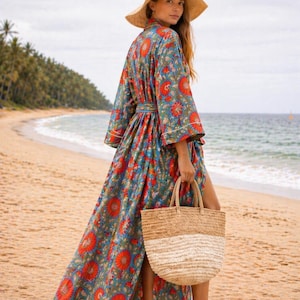 May include: A long, flowing floral print robe in shades of blue and red, worn with a straw hat and carried with a woven tote bag. The robe features a belt and white trim. The scene is set on a sandy beach with palm trees and ocean in the background.