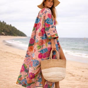 May include: A long, flowing pink floral print kimono-style robe with a straw hat and a woven tote bag. The robe features a vibrant pattern of large flowers in shades of pink, blue, and yellow. The tote bag is beige and white.