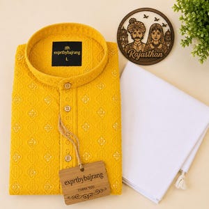 May include: A yellow embroidered kurta shirt with a mandarin collar and button-down front, paired with white pants. A wooden tag with the text "exprtbybajrang" hangs from the shirt. A wooden coaster with a couple illustration and the word "Rajasthan" is also visible.