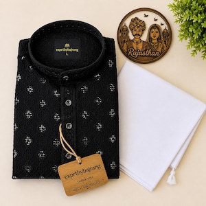 May include: A black long-sleeved shirt with a mandarin collar and a repeating geometric cutout pattern, featuring a front button placket and a wooden tag labeled exprtbybajrang. Beside it is a plain white folded fabric piece.