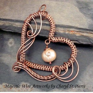 Może przedstawiać: Wisiorek w kształcie serca z drutu miedzianego z plecionym wzorem i pojedynczą perłą wiszącą w środku. Wisiorek znajduje się na ciemnoszarym tle.