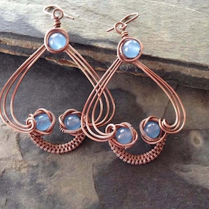 Puede incluir: Un par de pendientes de alambre de cobre con acentos de piedras preciosas azules. Los pendientes tienen forma de corazón con una forma de corazón más pequeña en la parte inferior. Las piedras preciosas están engastadas en el alambre y están rodeadas de alambre de cobre.