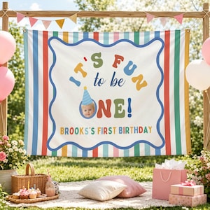 May include: A rectangular fabric banner featuring vertical stripes in pastel colors and a central white area with a wavy blue border. The text reads IT'S FUN to be ONE! with a photo of a child in a party hat and BROOKS'S FIRST BIRTHDAY.