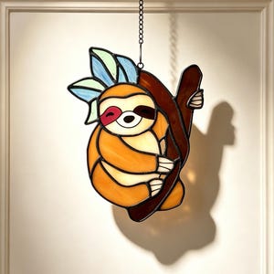 May include: A stained glass suncatcher depicting a sloth hanging from a dark brown tree branch. The sloth is crafted from orange and cream glass with dark brown eye patches. Above the sloth are light blue and green leaves, all suspended by a chain.