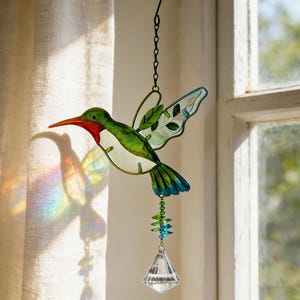 Puede incluir: Un atrapasoles de colibrí de vidriera con pico rojo, cabeza verde y cuerpo blanco. Las alas son verdes y azules, con un cristal transparente en la parte inferior. Cuelga de una cadena negra.