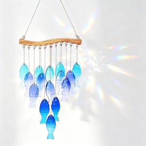Pode incluir: Um sino de vento decorativo com peças de vidro em forma de peixe em tons de azul. Os peixes estão suspensos de um galho de madeira natural com arame metálico. Ideal para decoração interior ou exterior, ambiente marítimo.