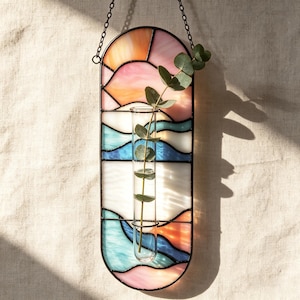 Può includere: Una decorazione da parete verticale in vetro colorato con parte superiore e inferiore arrotondate, caratterizzata da una tavolozza ispirata al tramonto con segmenti di vetro arancione, rosa, bianco e blu. Una provetta in vetro è fissata al centro.