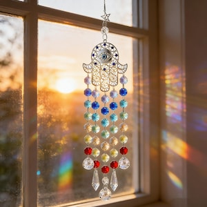 Può includere: Un acchiappasole decorativo con un design a mano di Hamsa argentata e un dettaglio a occhio diabolico. L'acchiappasole ha fili di perline sfaccettate colorate in un gradiente arcobaleno, con pendenti di cristallo trasparente in basso. È appeso davanti a una finestra.