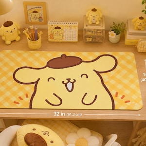 May include: A yellow desk mat with a cartoon character design, measuring 32 inches (81.3 cm) long and 16 inches (40.6 cm) wide. The mat features a smiling character with a brown beret on a yellow plaid background. Other desk accessories are visible.