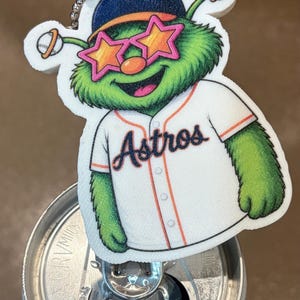 Puede incluir: Un llavero novedoso con un personaje de dibujos animados verde que lleva una camiseta de béisbol con la palabra "Astros" y gafas de sol en forma de estrella. El personaje lleva un sombrero azul y sostiene una pelota de béisbol.