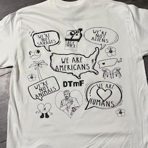 May include: White t-shirt with black graphic illustrations. The design includes speech bubbles with text like "We're not savages", "We are Americans", and "We are humans". Other illustrations include a camera, a frog, and a heart.