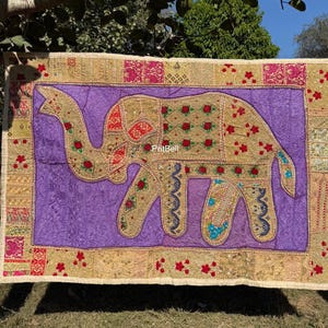 Op de afbeelding: Een rechthoekige wandtapijt met een olifantenontwerp. De olifant is goudkleurig met rode en groene bloemaccenten, tegen een paarse achtergrond. De rand heeft gouden, roze en rode patronen. De tekst "PritBell" is zichtbaar.