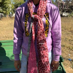 May include: A pink and red patterned scarf is draped over a person's neck, knotted in the front. The scarf has a repeating pattern of red shapes on a pink background. The person is wearing a purple hoodie and light blue jeans.