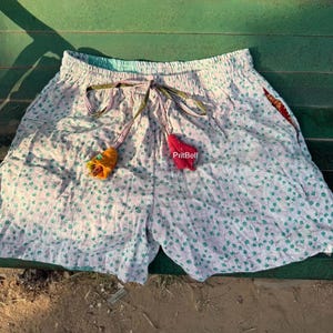 May include: Light-colored shorts featuring a small green leaf pattern, an elastic waistband with a drawstring, and two fabric tassels in yellow and red. The text PritBell is visible on the front. The shorts have a relaxed fit and a textured appearance.