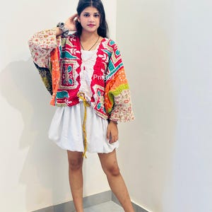May include: A multi-colored patchwork jacket featuring red, orange, and teal floral patterns with visible stitching, worn over a white gathered top with a drawstring waist and a white skirt. The text PritBell is visible on the jacket front.