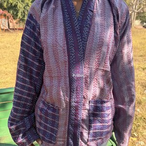 May include: A patchwork jacket in shades of purple, pink, and white. The jacket has a collared neckline, long sleeves, and two front pockets. The fabric has a textured appearance with visible stitching. The jacket is worn over a grey top.