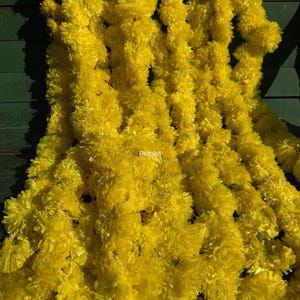 May include: A vibrant yellow garland with a fluffy, textured appearance. The garland is densely packed and appears to be made of a synthetic material. The background is a dark green surface. The word "Pritbell" is visible.