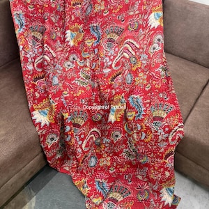 May include: A red, patterned quilt with a floral design. The quilt features a variety of colors, including blue, yellow, and white, on a red background. The quilt is draped over a brown sofa.