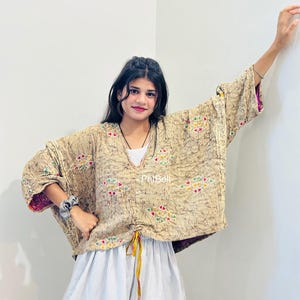 May include: A beige loose-fitting top with a dark brown crackle pattern, featuring a V-neckline and colorful geometric embroidery accents. The garment includes a drawstring hem with yellow and red ties, paired with white and blue striped shorts.