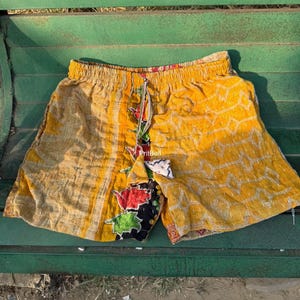 Può includere: Pantaloncini gialli con elastico in vita e chiusura con coulisse. Il tessuto presenta un aspetto trapuntato e testurizzato con un pannello verticale che mostra un motivo floreale in rosso, verde e nero. La parola PritBell è visibile.