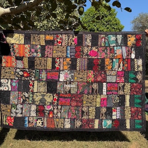 May include: A rectangular patchwork textile with a black border. The patchwork is composed of numerous rectangular pieces in various colors and patterns, including floral and geometric designs. The textile is held up outdoors.