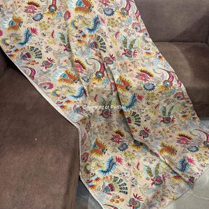 May include: A beige throw blanket with a colorful floral and paisley pattern. The design includes blue, yellow, red, and green accents. The blanket is draped over a brown sofa, showcasing its intricate details and soft texture.