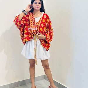 Peut inclure: Une veste de style kimono à motifs rouges et jaunes avec une fermeture à cordon sur le devant, portée sur un haut blanc et une jupe blanche à fines rayures verticales. La veste présente un imprimé géométrique et le texte PriBell est visible.