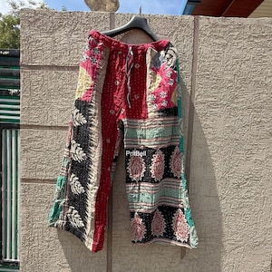 May include: Colorful patchwork pants with a drawstring waist. The pants feature a variety of patterns and colors, including red, black, pink, and green. The pants are hanging on a black hanger against a textured wall.
