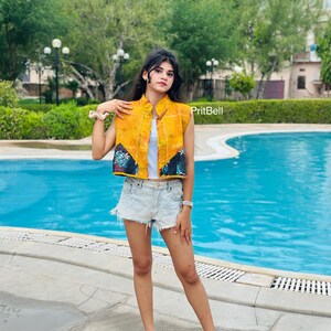 May include: A sleeveless yellow vest featuring a quilted texture and dark blue triangular panels with a floral pattern at the bottom hem. The garment is worn over a white top, paired with light blue denim shorts and black sandals.