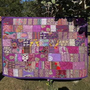 May include: A patchwork quilt with a vibrant array of purple hues. The quilt is composed of numerous fabric squares, each with unique patterns and textures. The edges are finished with a solid purple border. The quilt is displayed outdoors.