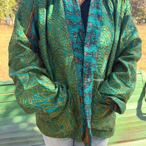 May include: A teal and gold patterned jacket featuring long sleeves, a wide collar, and two front pockets. The fabric displays an intricate abstract design with a distinct border pattern along the front opening. The text PritBell is visible.