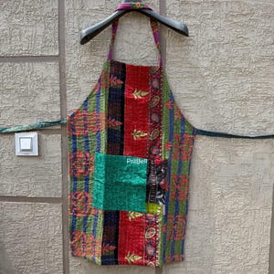 May include: A colorful apron with a patchwork design. The apron features a green pocket and is adorned with red, blue, and green patterns. The apron is hanging on a black hanger against a textured wall. The text "PritBell" is visible on the pocket.