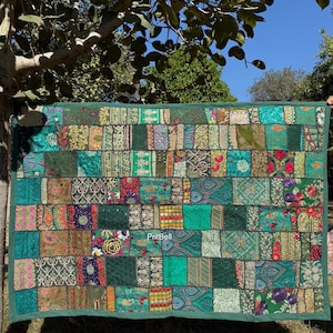 May include: A patchwork quilt with a variety of green fabrics, including teal, emerald, and olive tones. The quilt is rectangular and features a grid-like pattern of rectangular patches. The edges are finished with a solid green border. The quilt is hanging outdoors.