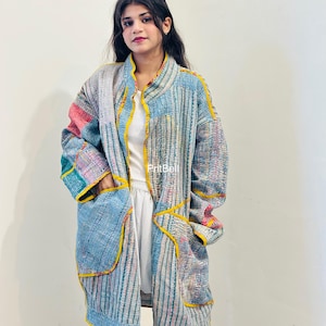 Könnte beinhalten: Eine langärmelige Jacke mit offener Vorderseite, die ein Patchwork-Design in Blau-, Grau- und Rosatönen aufweist, akzentuiert durch leuchtend gelbe Säume an den Kanten und Taschen. Das Kleidungsstück hat eine texturierte, gesteppte Optik.