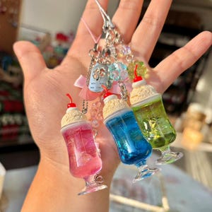 Könnte beinhalten: Eine Sammlung farbenfroher, miniaturisierter Milchshake-Anhänger. Jeder Anhänger zeigt ein klares Glas mit einer unterschiedlich gefärbten Flüssigkeit, gekrönt von einer Kugel Sahne und einer roten Kirsche. Die Anhänger sind an einem Schlüsselanhänger mit Perlen und einem kleinen Anhänger befestigt.
