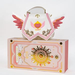 May include: A white handbag with pink and gold accents, featuring a heart and wing design. The bag has a clear handle and is placed on top of a pink box with gold celestial illustrations. The box has the text "U&D".
