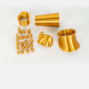 Puede incluir: Una colecci&oacute;n de accesorios de color dorado que incluye dos brazaletes anchos y curvos, una pieza cil&iacute;ndrica c&oacute;nica, un elemento decorativo con volantes y un conjunto de diecisiete componentes peque&ntilde;os c&oacute;nicos y cil&iacute;ndricos en cuadr&iacute;cula.
