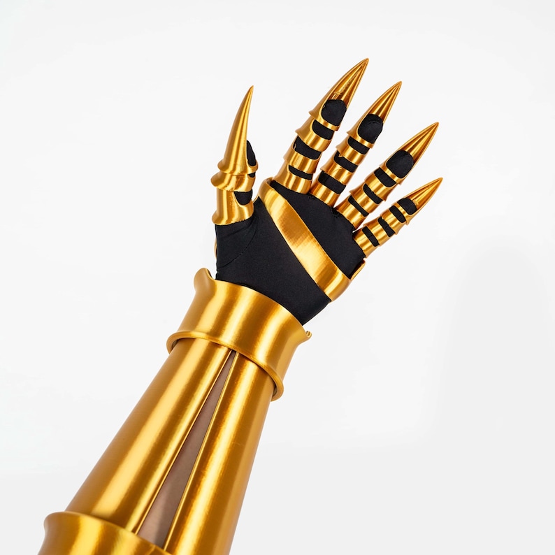 Puede incluir: Un guantelete de color dorado que presenta una placa larga para el antebrazo y una secci&oacute;n de mano con cubiertas articuladas para los dedos que terminan en puntas afiladas. La estructura se superpone sobre una base de tela negra con acabado met&aacute;lico.