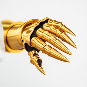 Puede incluir: Un guantelete de oro met&aacute;lico con segmentos de dedos articulados que terminan en puntas afiladas. La pieza de armadura est&aacute; construida con una capa base de tela negra visible en las articulaciones e incluye un pu&ntilde;o ancho y liso de color oro.
