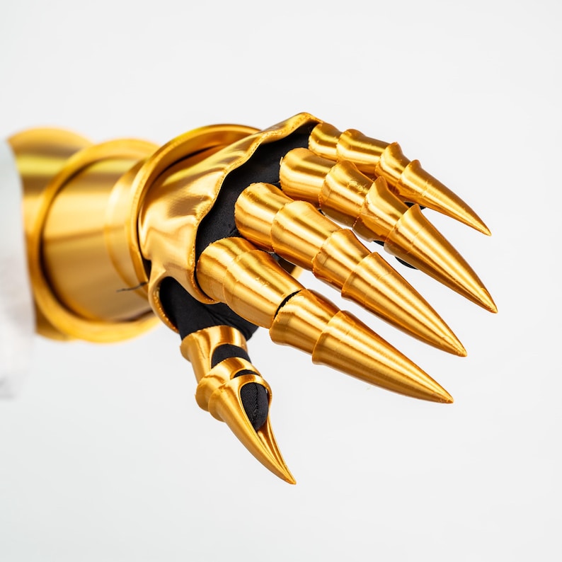 Puede incluir: Un guantelete de oro met&aacute;lico con segmentos de dedos articulados que terminan en puntas afiladas. La pieza de armadura est&aacute; construida con una capa base de tela negra visible en las articulaciones e incluye un pu&ntilde;o ancho y liso de color oro.