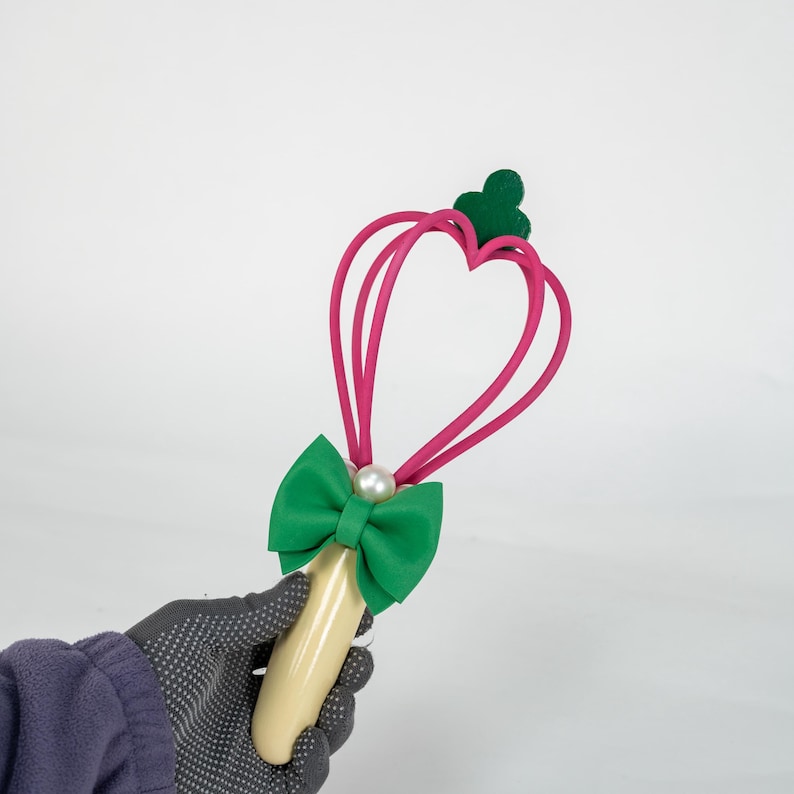 K&ouml;nnte beinhalten: Ein skurriler Zauberstab mit einem herzf&ouml;rmigen Design in Pink, einer gr&uuml;nen Schleife und einer Perle. Der Zauberstab hat einen cremefarbenen Griff. Die Oberseite des Zauberstabs hat ein gr&uuml;nes Kleeblatt-Design.